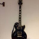 Černá elektrická kytara typu Les Paul se zlatým hardwarem a bílými blokovými vložkami na hmatníku. Na hlavě je viditelné logo Epiphone. Kytara je zav…