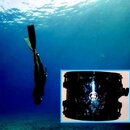 Fotografie zobrazuje potápěče v modré vodě a vložený detail černého cylindrického pouzdra z průhledného materiálu. Na přední straně pouzdra je vidite…