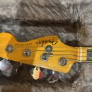 Fotografie zachycuje hlavu basové kytary z jasného dřeva s chromovanými ladičkami. Na dřevě je černý nápis Fender a vedle něj text JAZZ BASS. Nástroj…