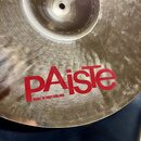 Detailní pohled na kovový činel zlatavé barvy s viditelnou strukturou broušení. Na povrchu je červený nápis PAiste a pod ním menší text MADE IN SWITZ…