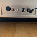 Detail zadní strany stříbrného zařízení s konektory. Jsou zde nápisy MIDI-OUT, R OUT L, HEADPHONES a DC 12V. Do napájecího konektoru je zapojen černý…