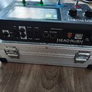 Černý procesor efektů HeadRush Gigboard umístěný na stříbrném hliníkovém kufru s kovovými zapínacími zámky. Na zadním panelu jsou viditelné nápisy PA…