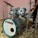 Modrá bicí souprava s kovovým hardwarem. Na předním koži basového bubnu je černý nápis SONOR s doprovodným logem. Na jednom z tomů je viditelný malý…
