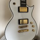 Bílá elektrická kytara s černým lemováním a zlatým hardwarem. Nástroj má dva humbuckery se zlatými kryty, Tune-o-matic kobylku a stopbar strunovník.…