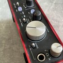 Detailní pohled na horní panel červeného audio rozhraní s černým povrchem. Jsou vidět stříbrné a černé ovratové ovladače s nápisy GAIN, MONITOR a SOU…