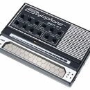 Černý přenosný syntetizátor s bílým nápisem Stylophone TM GEN X-1. Horní panel obsahuje řadu ovládacích knoflíků s popisky jako ATTACK, PITCH, DECAY,…
