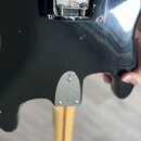 Detail zadní strany černé elektrické kytary. Je vidět kovový mechanismus pružin kobylky a kovová krytka u základny krku připevněná šrouby. Povrch lak…