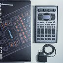 Na fotografii je sampler Roland SP-404MKII v šedé barvě s mřížkou čtvercových padů. Vedle přístroje je umístěna originální krabice s nápisy Roland a…