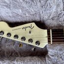 Fotografie zachycuje hlavu elektrické kytary v krémové barvě. Na povrchu je černý nápis Fender a STRATOCASTER. Nástroj má šest chromovaných ladiček a…