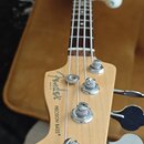 Fotografie zachycuje hlavu a hmatník elektrické basové kytary z přírodního světlého dřeva s tmavým hmatníkem. Na hlavě je černé logo Fender a pod ním…