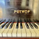 Detail klaviatury a horní části černého pianina. Na lakovaném povrchu je zlatým nápisem uvedena značka PETROF, v okolení nápisu jsou vidět četné škrá…