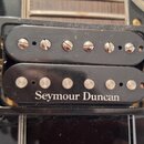 Fotografie zachycuje dva elektrické snímače typu humbucker v černém plastovém rámečku. Spodní snímač nese bílý nápis Seymour Duncan a má viditelné šr…