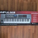 Fotografie znázorňuje krabici s MIDI klaviaturou v barvě stříbrné a černé. Na krabici jsou nápisy IMPULSE 61, novation a text 61 NOTE PROFESSIONAL US…