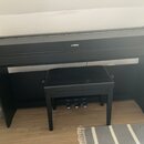 Černý digitální piano s přívodním stolem a černou polstrovanou stolečkou. Na přední horní hraně je bílé logo a nápis YAMAHA. Nástroj má tři stupně pe…