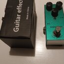 Zelený efekt pedál s nápisy Analog Delay a TRUE BYPASS. Na horní straně jsou tři černé ovládací knoflíky a stisknutý spínač. Vedle pedálu je černá ka…