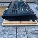 Černý mixovací pult s velkým množstvím modrých a zelených posuvníků. Na přední hraně je bílý nápis: "MIXING CONSOLE 16 x 2 + 2 MIXING CONSOLE INTERNA…