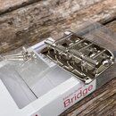 Kovový stříbrný kobylka (bridge) pro elektrickou kytaru s šesti sedly a montážními šrouby. Nástroj je umístěn v plastovém obalu na bílém kartonovém b…