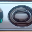 Fotografie znázorňuje otevřené bílé balení s dvěma kabely. Vlevo je krátký tyrkysový kabel s USB konektorem, vpravo je srolovaný černý kabel. Na před…