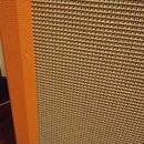 Detailní záběr na roh reproduktorové skříně s oranžovým povrchem. Přední strana je kryta béžovou tkanou síťovkou. Na oranžovém okraji je viditelný dr…