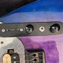 Detailní pohled na ovládací panel elektrické kytary s modrofialovým přechodem barev. Černý plastový panel obsahuje dva otočné knofíky a jeden přepína…