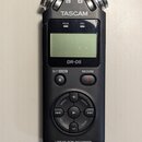 Černý digitální záznamový zařízení s nápisem TASCAM a označením modelu DR-05. Na horní části jsou dvě stříbrná mikrofonní kapsle s označením L a R. P…