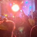 Fotografie zachycuje koncertní scénu s osvětlením a davem lidí v popředí. Na pozadí je vidět kovová konstrukce pódia a jasný zdroj světla. V pravém h…