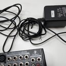 Černý síťový adaptér značky BEHRINGER s nápisem MODEL: SMPS-12V. Na boku je nalepený bílý štítek s číslem 209. Vedle adaptéru je umístěn horní panel…