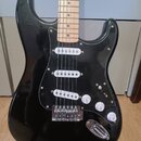 Černá elektrická kytara typu Stratocaster se světlým javorovým hmatníkem. Má černý pickguard a tři bílé snímače typu single-coil. Na ovládacích knofl…