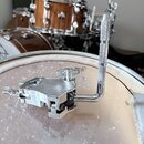 Stříbrný kovový pedál na bicí položený na bílém bubnu. Nástroj nemá žádné viditelné značky ani nápisy. Povrch je lesklý a v dobrém stavu. V pozadí js…