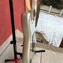Fotografie zachycuje zadní část elektrické kytary v šampaňské nebo zlatavé barvě, která stojí na černém stojanu. Na svislé tyči stojanu je bílý nápis…