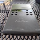 Šedý obdélníkový elektronický přístroj s nápisy KAWAI a Pocketband. Horní část obsahuje malý displej, několik kulatých tlačítek a texty jako PHY VOL,…