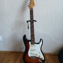 Elektrická kytara s trupem v barevnosti sunburst a bílým pickguardem, umístěná na černém stojanu. Na hlavě hmatníku je viditelné logo Fender. Nástroj…