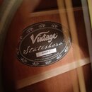 Fotografie zachycuje vnitřní štít akustické kytary přilepený k dřevěnému korpusu. Černý oválný štít nese bílé nápisy „Vintage“ a „Stateshora'“. Pod n…