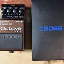 Kytarový efekt pedál značky BOSS, model Octave OC-5 v hnědé barvě. Na pedálu jsou nápisy OUTPUT, DIRECT OUT a INPUT. Vedle pedálu je umístěna černá k…