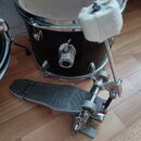 Černý buben s kovovými obručemi a bílým koženem. Před ním je umístěn kovový pedál s nápisem CB DRUMS na stupni. K pedálu je připojena kovová tyč s bí…