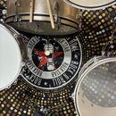 Fotografie zachycuje část bicí sady v zlaté barvě s kovovými obručemi. Na přední kože bass drumu je černobílý grafický printed s červenou hvězdou a n…