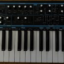 Černý analogový syntetizátor s bílými a černými klávesami. Na levé straně je nápis novation BASS STATION 2 Analogue synthesizer. Přední panel obsahuj…