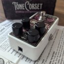 Bílý efekt pedál s nápisem Tone Corset a fialovým ornamentálním vzorem na horní ploše. Pedál má čtyři černé ovládací knoflíky a jeden stomeč. V pozad…