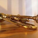 Mosazný trumpet v zlaté barvě položený na dřevěném stole. Nástroj vykazuje známky opotřebení povrchu a oxidaci v několika místech. Na fotce nejsou žá…