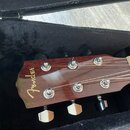 Detail hlavy akustické kytary z tmavého dřeva v černém pouzdře. Na hlavě je viditelné zlaté logo Fender. Jsou zde vidět šest kovových ladiček a strun…