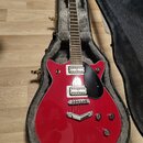 Červená elektrická kytara s černým pickguardem v otevřeném kufru s šedou vnitřní výstuhou. Na hlavě hmatníku je bílý nápis Gretsch. Kytara má dva hum…