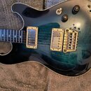 Elektrická kytara s tmavě modrým průsvitným lakem, pod kterým je vidět struktura dřeva. Nástroj má dva humbuckery s krémovými rámečky, jeden tónový p…