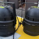 Dvě černé reflektory v kovovém provedení s horními madly. Na horní části obou zařízení jsou žluté samolepky s nečitelným textem. Levý reflektor má na…