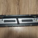 Fotografie zachycuje vnitřní mechanismus hudebního přístroje v černém plastovém šasi. Uprostřed je umístěna hliníková lišta s dvěma paralelními kovov…