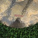 Detailní pohled na kovový činel s kovaným povrchem. Na povrchu je červený nápis "Troy" a pod ním logo s nečitelným textem. Činely vykazují mírné oxid…