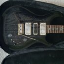 Elektrická kytara s tmavým, pruhovaným povrchem těla v černém kufru. Nástroj má tři bílé humbuckery, stříbrný most a čtyři ovládací knoflíky. Na hmat…