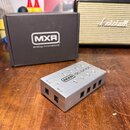 Stříbrný kovový napájecí adaptér s nápisem MXR dc brick. Na boku jsou čtyři výstupy, dva z nich mají popisy 10V out a 18V out. Vedle zařízení je čern…