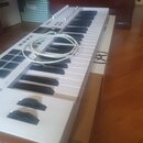 Bílá klávesová syntezátor s černými klávesami a několika ovládacími prvky v horní části. Na přístroji je položen bílý kabel. Povrch nástroje vypadá č…