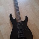 Černá elektrická kytara s tmavým hnedým hmatníkem a dvěma humbuckery. Na hmatníku je viditelný bílý štítek s nápisem "No 107". Na hlavě krku je bílé…