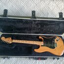 Elektrická kytara s přírodním dřevěným povrchem těla a hmatníkem, umístěná v černém polstrovaném kufru. Na hlavě je vidět nápis Fender STRATOCASTER.…